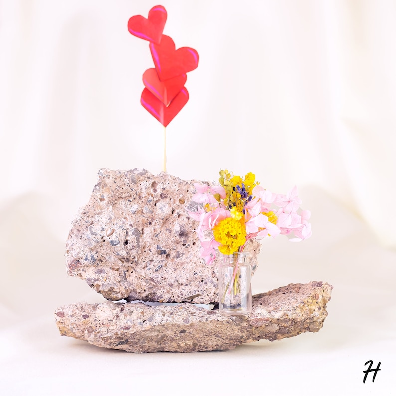 May include: A small glass vase filled with colorful dried flowers, including yellow and pink blooms, sits on a piece of rough, gray stone. Above, a wooden stick holds three red paper hearts. The arrangement is set against a white backdrop.