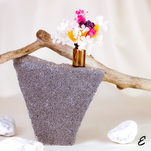 May include: A small brown glass bottle filled with colorful dried flowers, including white, yellow, pink, and purple blooms. The bottle sits atop a gray, triangular concrete block, with a wooden branch and white stones in the background.