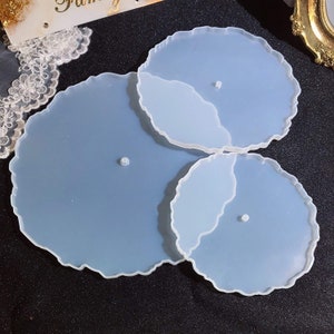 May include: A set of three resin coasters in different sizes, featuring an irregular, organic design. The coasters are a gradient of light and medium blue, with a white edge. Each coaster has a small, circular foot.