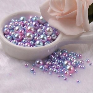 Puede incluir: Un conjunto de perlas iridiscentes en tonos rosa, morado y azul, desbordando de un cuenco de cer&aacute;mica blanco. Una suave rosa rosa descansa junto a las perlas, creando una exhibici&oacute;n delicada y elegante.