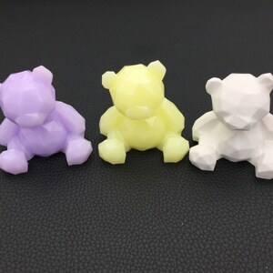 May include: Three geometric bear figurines in lavender, yellow, and white. Each bear is sitting with its legs forward and has a faceted design. The bears are displayed on a dark surface.
