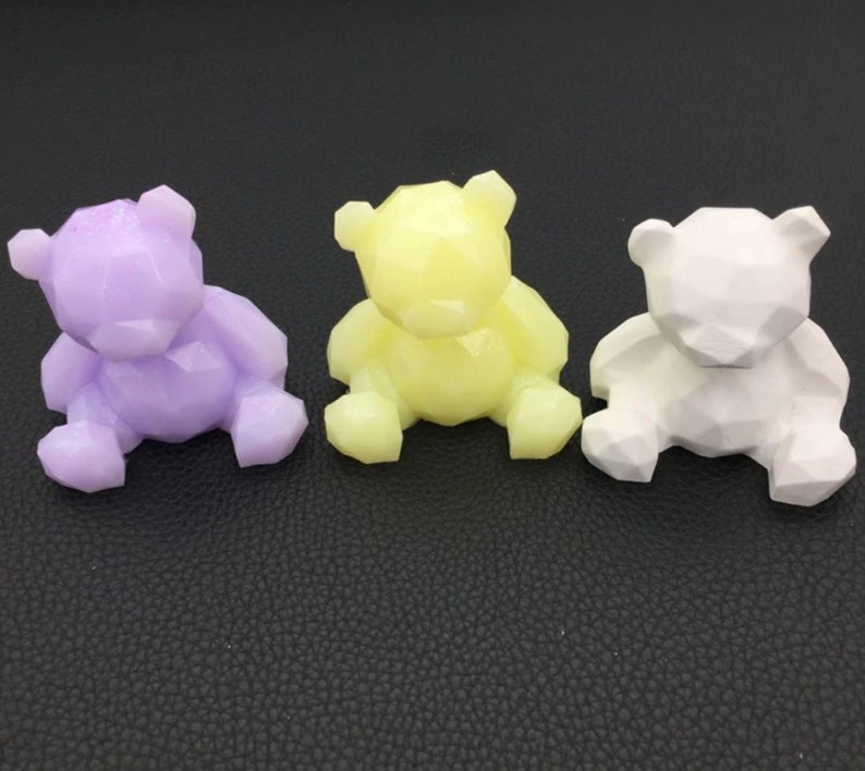 May include: Three geometric bear figurines in lavender, yellow, and white. Each bear is sitting with its legs forward and has a faceted design. The bears are displayed on a dark surface.