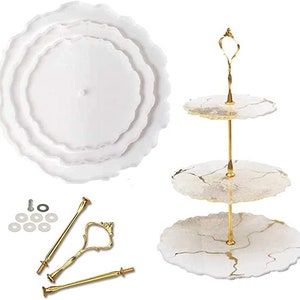 May include: A white, three-tiered serving tray with gold-coloured hardware. The tray has a wavy edge design. The hardware includes a handle and connecting rods. Also included are the components to assemble the tray.