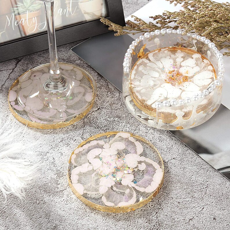 May include: Clear resin coasters with a floral design, gold leaf accents, and a gold rim. One coaster is under a wine glass. A matching coaster holder with pearl trim holds a coaster. The coasters are decorative and functional.