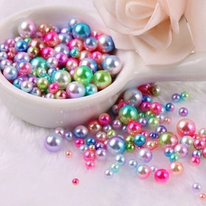 Puede incluir: Una colecci&oacute;n de perlas de imitaci&oacute;n iridiscentes en varios tama&ntilde;os y colores, incluyendo rosa, verde, azul y morado, se exhiben en un cuenco de cer&aacute;mica blanco. Algunas perlas est&aacute;n esparcidas sobre una superficie blanca y esponjosa, con una flor rosa suave en el fondo.