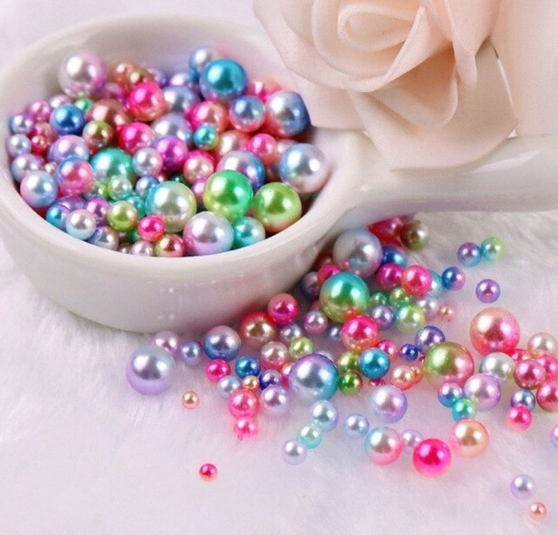 Puede incluir: Una colecci&oacute;n de perlas de imitaci&oacute;n iridiscentes en varios tama&ntilde;os y colores, incluyendo rosa, verde, azul y morado, se exhiben en un cuenco de cer&aacute;mica blanco. Algunas perlas est&aacute;n esparcidas sobre una superficie blanca y esponjosa, con una flor rosa suave en el fondo.