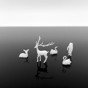 May include: A monochrome image showing small white figurines of a deer, a fawn, two swans, and a person in a winter coat. The figurines are set on a reflective black surface, with a gradient white background.