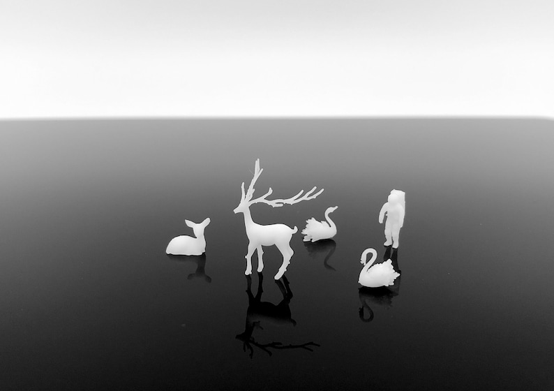 May include: A monochrome image showing small white figurines of a deer, a fawn, two swans, and a person in a winter coat. The figurines are set on a reflective black surface, with a gradient white background.