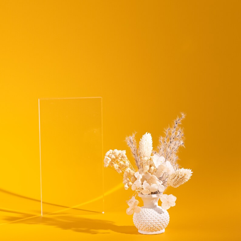 May include: A small white vase with a textured surface holds a bouquet of dried flowers in shades of cream and beige. A clear, rectangular acrylic panel stands to the left against a bright yellow background. The scene is lit with strong directional light.