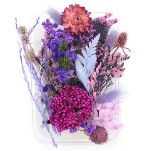 May include: A collection of dried flowers in shades of purple, pink, and magenta, including a large strawflower, sprigs of lavender, small purple statice, a light blue fern leaf, pink baby's breath, and fluffy purple bunny tail grass.
