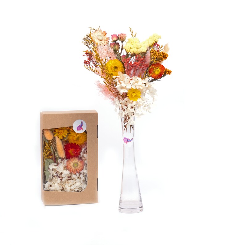 Peut inclure: Un vase en verre transparent contient un bouquet de fleurs s&eacute;ch&eacute;es aux tons jaunes, oranges, roses et blancs. Une bo&icirc;te en carton avec une fen&ecirc;tre pr&eacute;sente un arrangement similaire de fleurs s&eacute;ch&eacute;es. Un autocollant papillon violet est sur le vase et la bo&icirc;te.