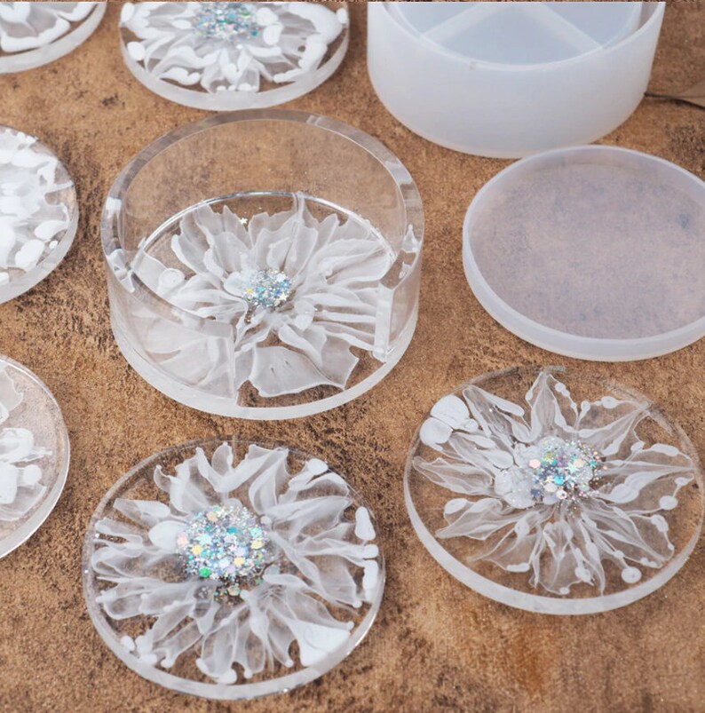 May include: Clear resin coasters and a coaster holder with white floral designs and glitter accents. The coasters are round, and the holder is cylindrical. The designs are centered with a glittery embellishment.