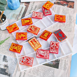 May include: A collection of rectangular, resin-cast objects with intricate designs in red, orange, and gold. The objects are arranged in a white, grid-like tray, suggesting a crafting or organizational context. The designs appear to be illustrations.