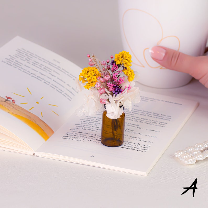 May include: A small brown glass bottle filled with colorful dried flowers, including yellow, pink, and white blooms. The bottle sits on an open book with text, next to a white mug and a pearl hair clip.
