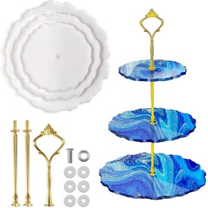 May include: A white, three-tiered serving tray with scalloped edges and a gold-coloured handle. The tray is shown disassembled with the hardware. A separate three-tiered tray with blue and white marbled design is also shown.