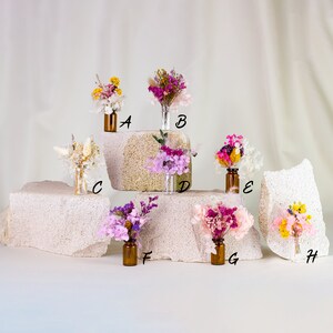 May include: Assortment of dried flower bouquets in small glass bottles. The bouquets feature various colors, including yellow, pink, purple, and white. The bottles are arranged on textured, off-white blocks against a neutral background.