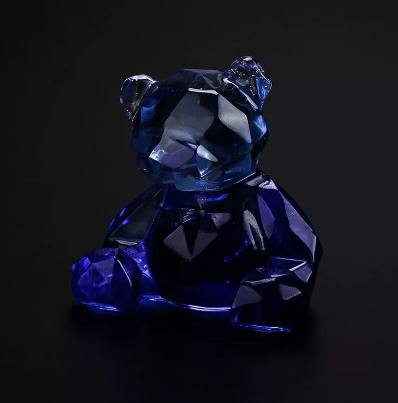 May include: A faceted, blue crystal bear figurine. The bear is sitting with its legs forward and its arms at its sides. The crystal is a deep blue color and has many facets that reflect light. The background is black.