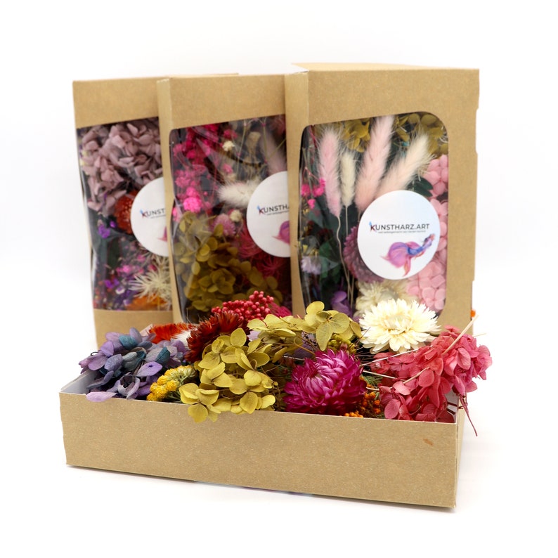 Peut inclure: Trois bo&icirc;tes en papier kraft avec des fen&ecirc;tres transparentes remplies de fleurs s&eacute;ch&eacute;es et une bo&icirc;te rectangulaire de fleurs s&eacute;ch&eacute;es de diff&eacute;rentes couleurs. Les bo&icirc;tes ont un autocollant avec l'inscription "KUNSTHARZ.ART". Les fleurs sont roses, violettes, jaunes et rouges.