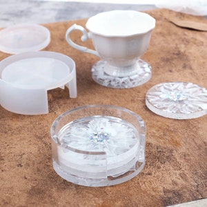 May include: A clear resin coaster set with a white flower design and glitter accents. The set includes a holder, individual coasters, and a teacup. The coasters are approximately 4 inches in diameter.