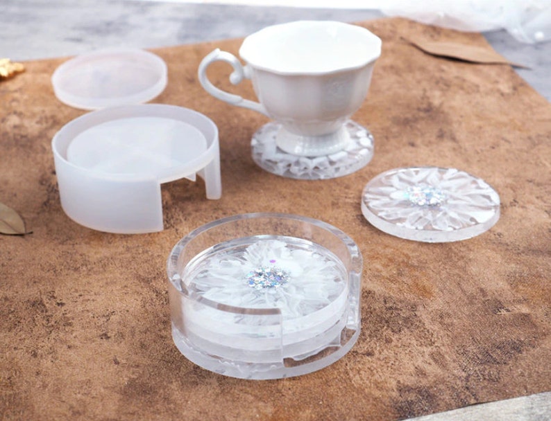 May include: A clear resin coaster set with a white flower design and glitter accents. The set includes a holder, individual coasters, and a teacup. The coasters are approximately 4 inches in diameter.