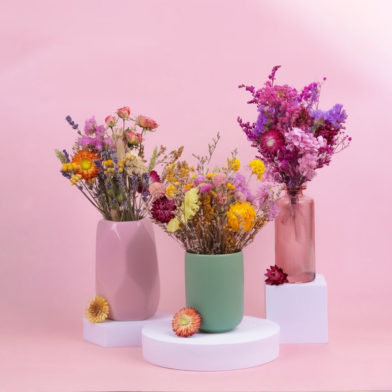 Peut inclure: Trois vases de fleurs s&eacute;ch&eacute;es dans des tons roses, verts et en verre transparent. Les fleurs sont un m&eacute;lange de couleurs, dont le rose, le violet, le jaune et l'orange. Les vases sont pos&eacute;s sur des socles blancs sur fond rose.