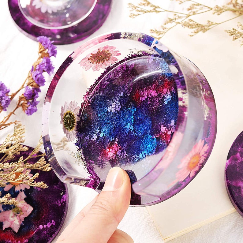 May include: Clear resin coasters with embedded dried flowers and a galaxy-inspired design. The coasters feature a mix of purple, blue, and pink hues, with a glossy finish. The set includes multiple coasters, perfect for home decor.