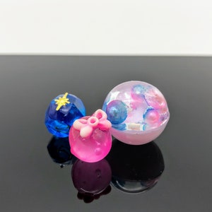 Puede incluir: Tres objetos decorativos de resina: una gema azul facetada con una estrella dorada, un orbe rosa con un lazo y una esfera transparente llena de orbes coloridos. Los objetos se exhiben sobre una superficie negra reflectante.