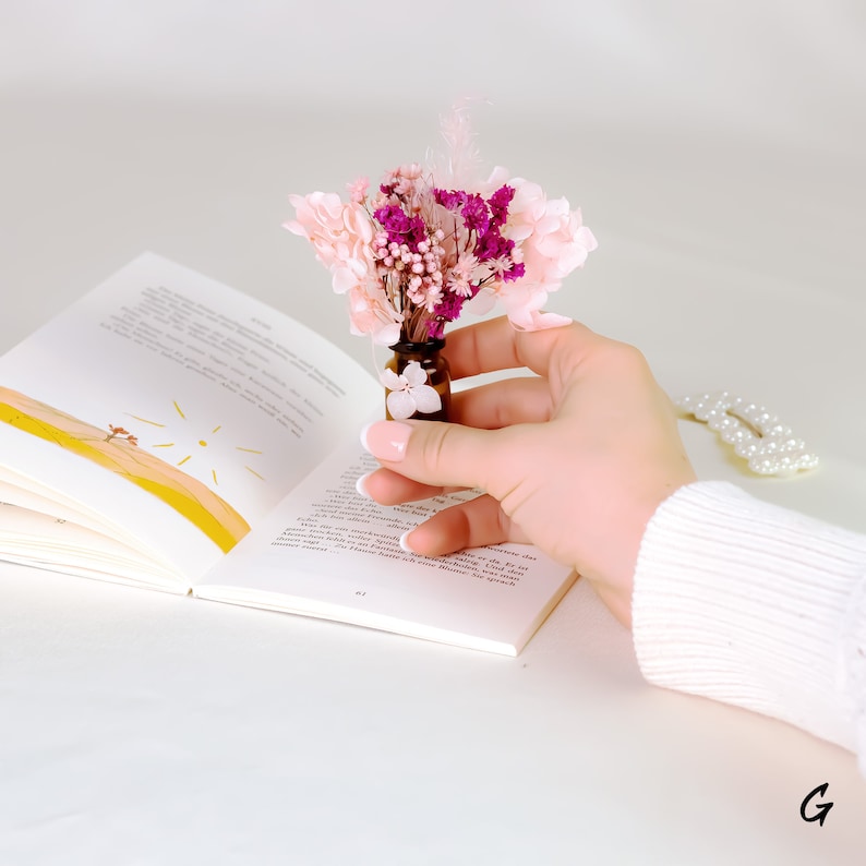 May include: A small brown glass bottle filled with dried pink and purple flowers. The bottle is held over an open book with illustrations. A white pearl hair clip rests on the surface.