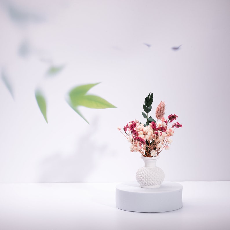 May include: A small white vase with a textured surface holds a bouquet of dried flowers in shades of pink, red, and green. The vase sits on a white cylindrical pedestal against a soft white background with blurred green leaf shadows.