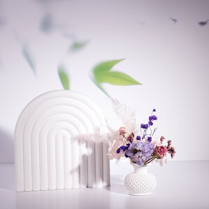 May include: A white, arched decorative piece with a series of concentric semi-circles. A small white vase with a textured pattern holds a bouquet of dried flowers in shades of purple, pink, and cream. The background is a soft white with blurred green leaf shapes.