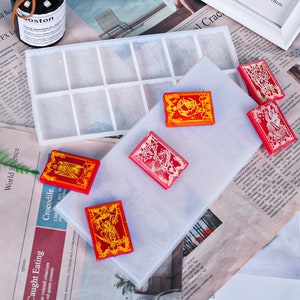 May include: A collection of red and gold resin card-shaped objects with intricate designs. A clear rectangular mold with multiple compartments is visible, along with a clear sheet. The cards have detailed patterns and are arranged on a newspaper background.