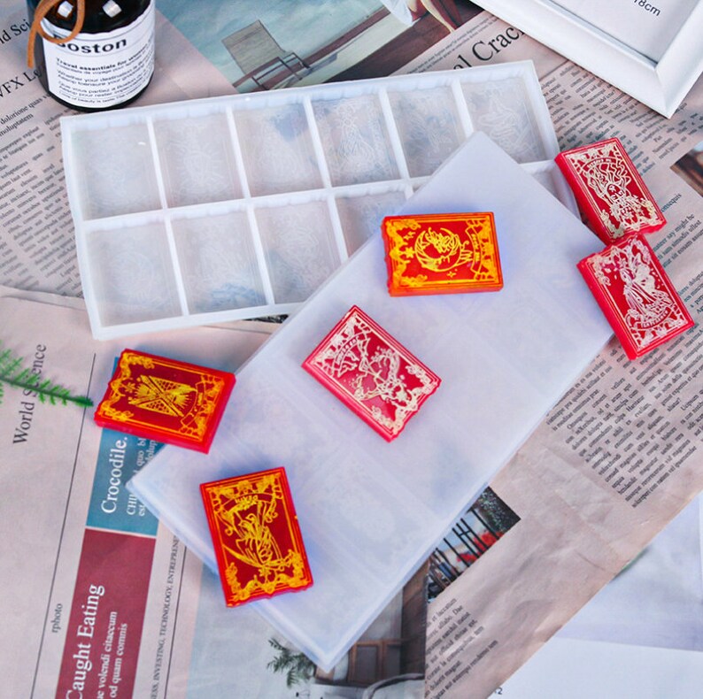 May include: A collection of red and gold resin card-shaped objects with intricate designs. A clear rectangular mold with multiple compartments is visible, along with a clear sheet. The cards have detailed patterns and are arranged on a newspaper background.