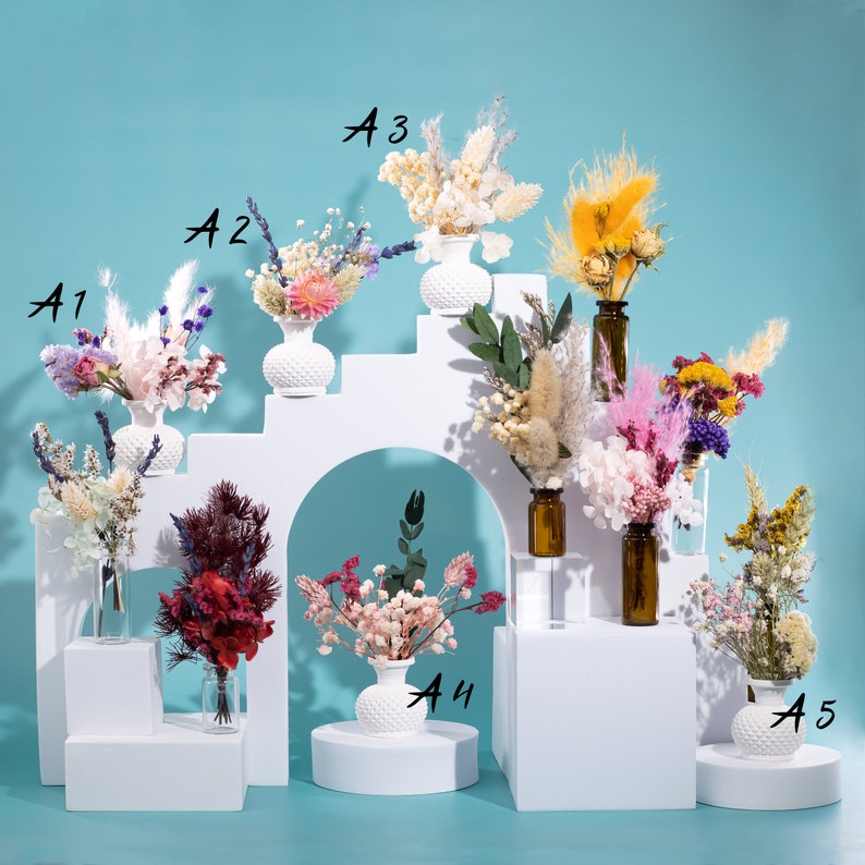 May include: Arrangement of dried flowers in small vases and bottles. The flowers are in various colors, including pink, purple, yellow, and white. The vases are white and have a textured surface. The bottles are amber-colored.