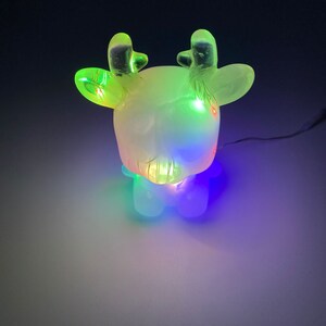 May include: A glowing reindeer figurine with a frosted body and clear antlers. The reindeer is illuminated by colorful LED lights, creating a festive and whimsical display. The lights cycle through green, blue, and other colors.