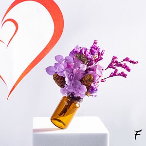 May include: A small amber glass bottle filled with a bouquet of purple and lavender flowers, including hydrangeas and dried sprigs. The bottle is tilted on a white block, with a red heart outline in the background.