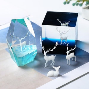 May include: Two decorative resin sculptures featuring white deer figures. One is a blue and clear geometric shape, the other a black and blue cube. Several white deer figurines are also present.