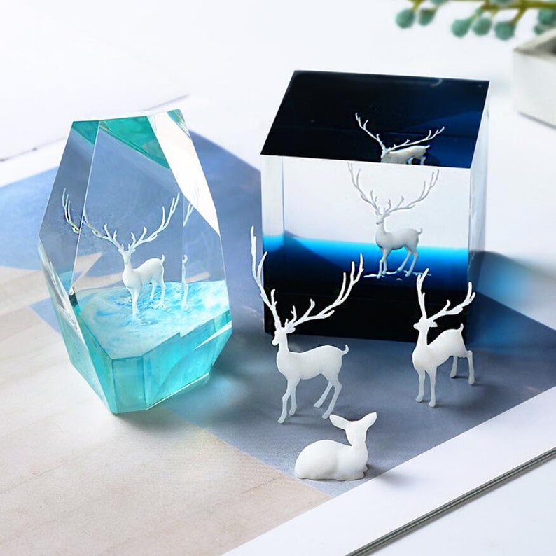 May include: Two decorative resin sculptures featuring white deer figures. One is a blue and clear geometric shape, the other a black and blue cube. Several white deer figurines are also present.