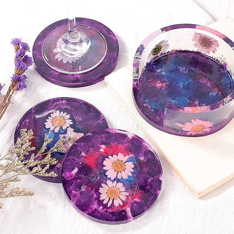May include: A set of purple resin coasters with embedded dried flowers. The coasters have a galaxy-like design with white and pink daisy flowers. Includes a coaster holder and a coaster with a wine glass.