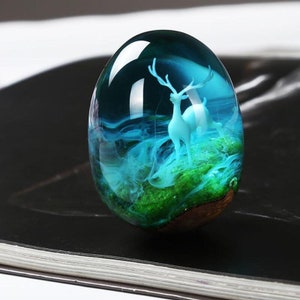 May include: An egg-shaped resin ornament with a white deer figurine inside a miniature landscape. The scene includes green moss and blue, swirling, cloud-like formations, all encased in the transparent resin. The base is a dark wood.