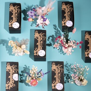 May include: Arrangement of dried flowers in small white vases and black boxes with gold floral designs. Each box has a sticker with a name on it, such as "Anna" and "Maria". The background is a light blue.