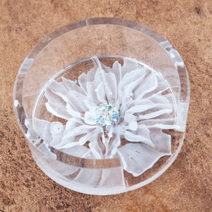 May include: A clear, round coaster with a white floral design. The flower has delicate petals and a sparkling, glittery center. The coaster is made of a transparent material, allowing the design to be visible.