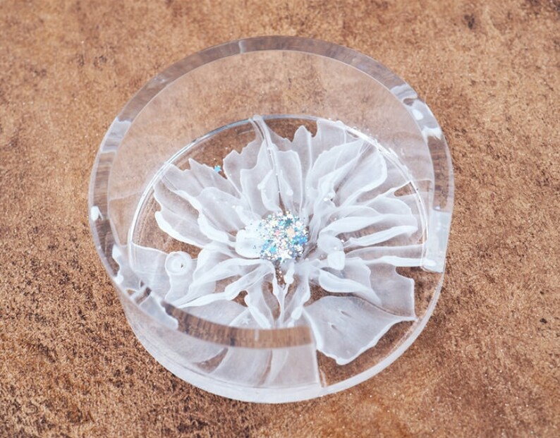 May include: A clear, round coaster with a white floral design. The flower has delicate petals and a sparkling, glittery center. The coaster is made of a transparent material, allowing the design to be visible.