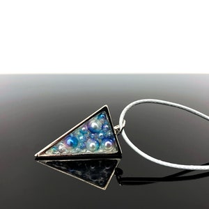 Puede incluir: Un collar con colgante triangular plateado. El colgante est&aacute; relleno de cuentas azules y blancas de varios tama&ntilde;os, creando una apariencia texturizada. El collar tiene un cord&oacute;n blanco.