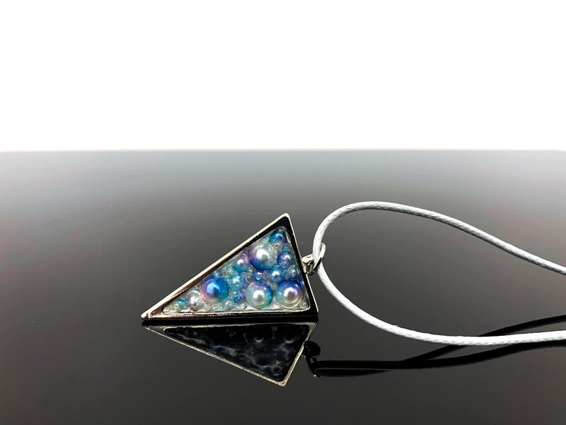 Puede incluir: Un collar con colgante triangular plateado. El colgante est&aacute; relleno de cuentas azules y blancas de varios tama&ntilde;os, creando una apariencia texturizada. El collar tiene un cord&oacute;n blanco.