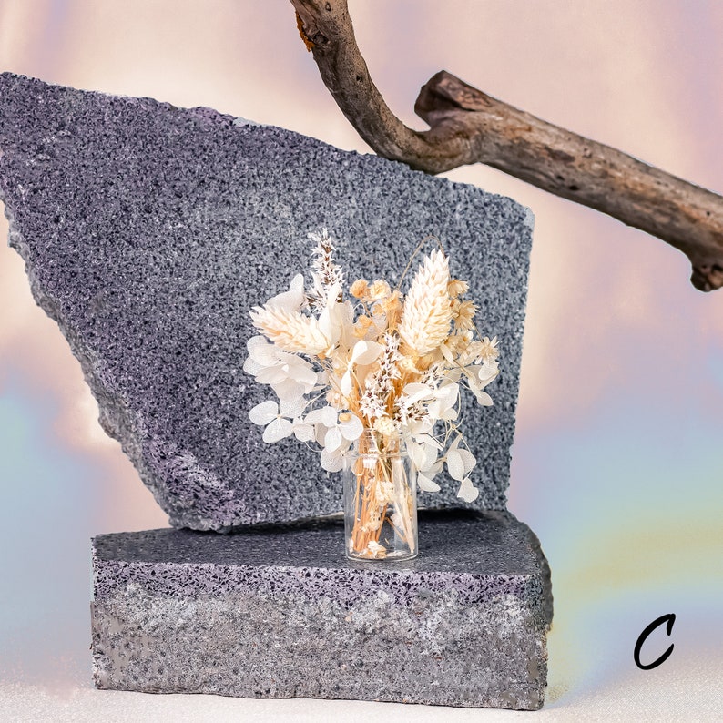 May include: A small glass vase filled with dried flowers in shades of white and beige sits on a gray, textured block. A second gray block and a piece of driftwood are in the background. The letter "C" is in the lower right corner.