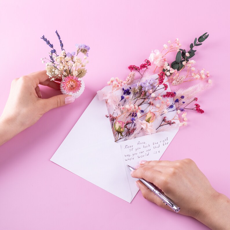 May include: A white envelope with dried flowers and a handwritten note that reads "Dear Anna, If you look the right way, you can see that the whole world is a..." A small floral arrangement is held in the hand.