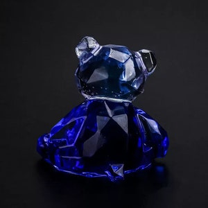 May include: A faceted, dark blue glass bear figurine. The bear is sitting, with its head turned slightly to the side. The glass has a crystalline appearance, catching the light and creating a sparkling effect. The bear is a decorative item.