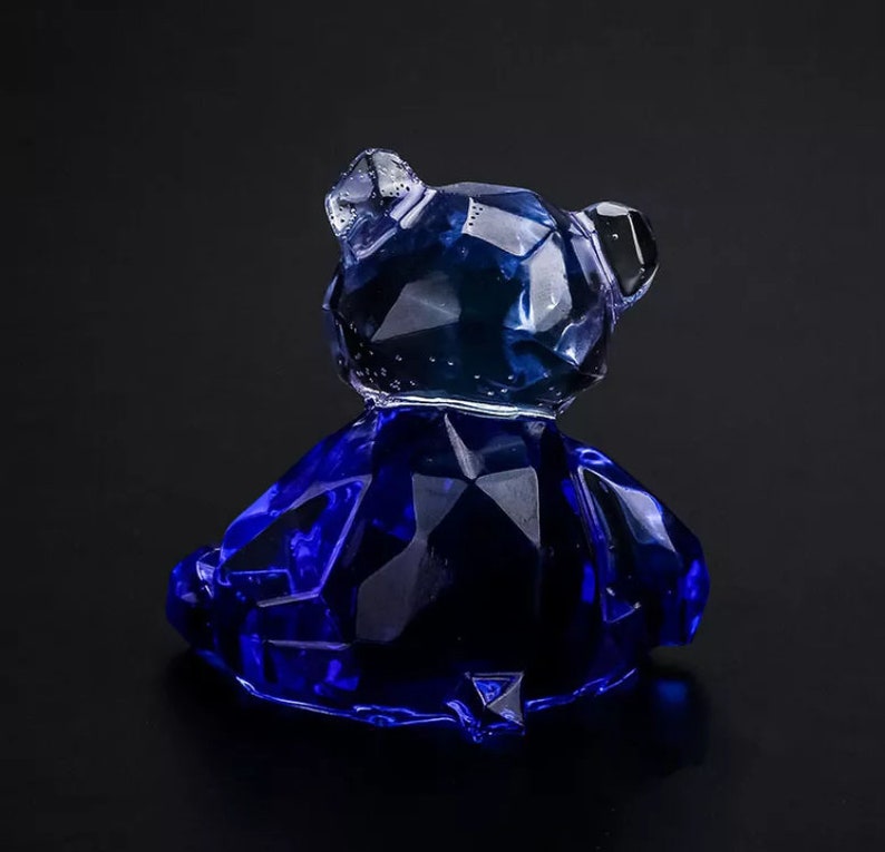 May include: A faceted, dark blue glass bear figurine. The bear is sitting, with its head turned slightly to the side. The glass has a crystalline appearance, catching the light and creating a sparkling effect. The bear is a decorative item.