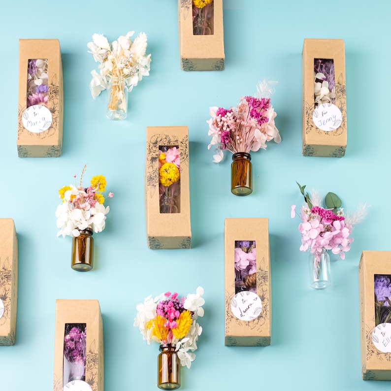 May include: Arrangement of small glass bottles filled with dried flowers in various colors, including pink, yellow, and white. Some bottles are paired with rectangular cardboard boxes with a window, and some have handwritten labels.