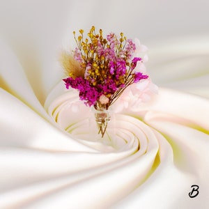 May include: A small glass vase filled with a bouquet of dried flowers. The arrangement includes vibrant magenta, yellow, and pale pink blooms, with a touch of beige. The vase sits on a cream-colored, draped fabric.